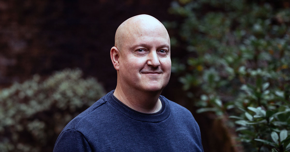 Mike Mingard standing outdoors, smiling slightly with a blurred natural background.