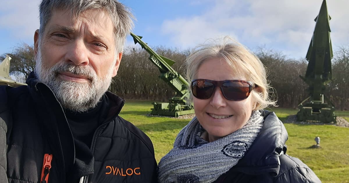 Bjørn, wearing a Dyalog-branded jacket, stands beside a smiling woman in sunglasses and a scarf. Behind them are two large green military missiles on display in an outdoor museum or exhibit area under a blue sky.