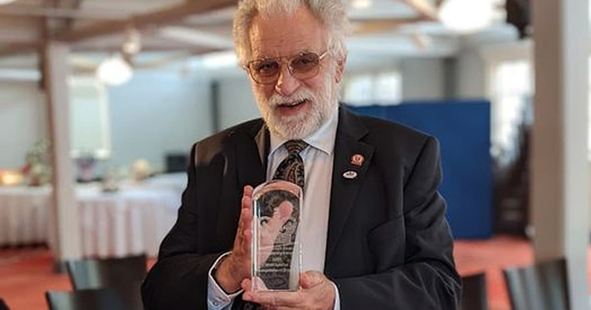 Geoff Streeter holding an award trophy, dressed in a dark suit and patterned tie, smiling at the camera in a warmly lit room.