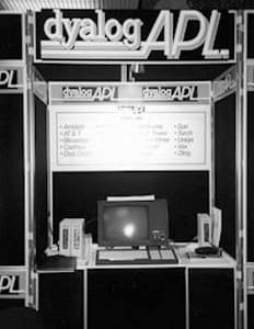 Black and white photo of an early Dyalog APL exhibition booth, featuring a CRT monitor, keyboard, manuals, and large signage with the Dyalog APL logo.