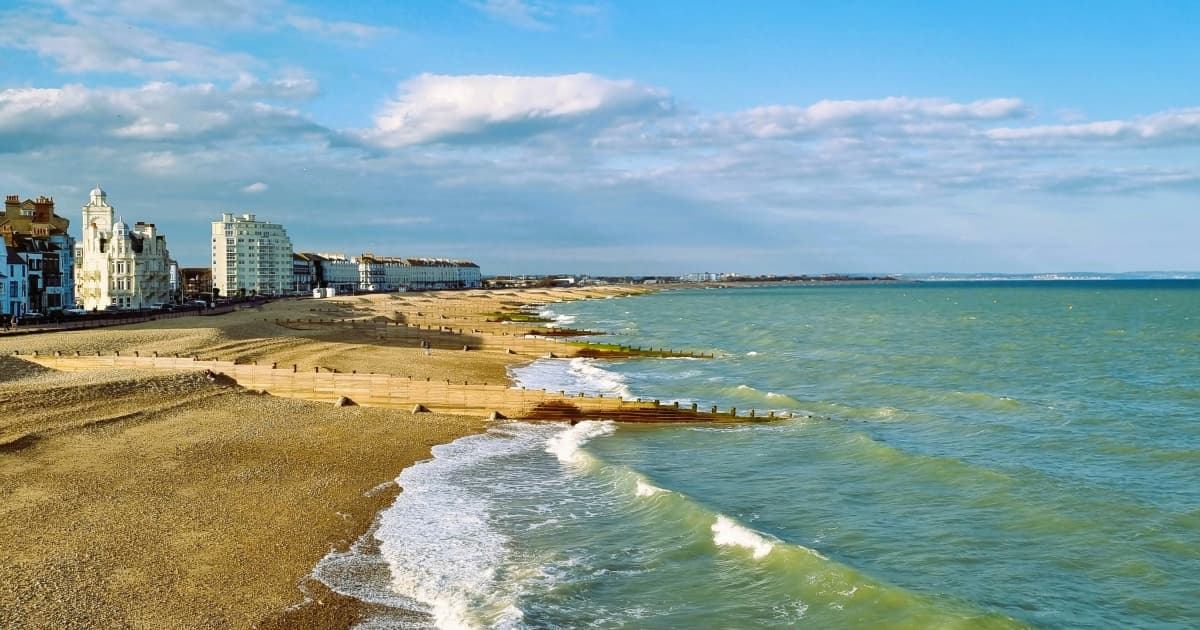 A wide view of Eastbourne seafront with waves breaking on the pebble beach, wooden groynes stretching into the sea, and a line of buildings including hotels and apartments along the promenade under a partly cloudy sky.