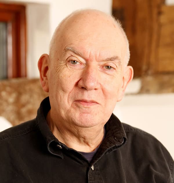 John Scholes in a warmly lit room, wearing a dark shirt and looking directly at the camera with a calm expression.