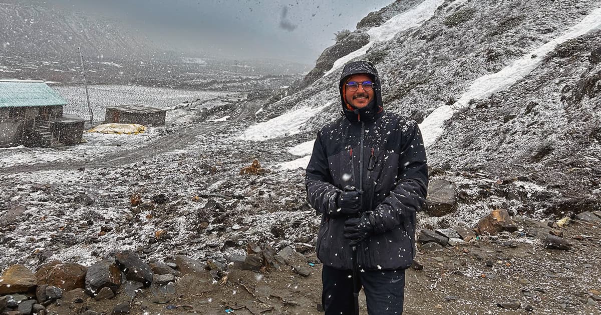 Aarush Bhat standing in a snowy mountain landscape, wearing a black winter jacket and holding a walking pole.