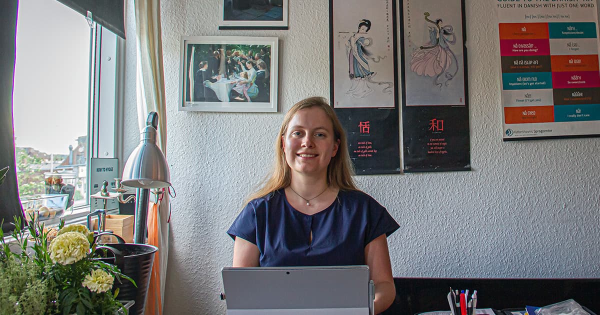 Andrea Plovgaard Frederiksen working at her desk in a bright, plant-filled room decorated with cultural posters and framed art.