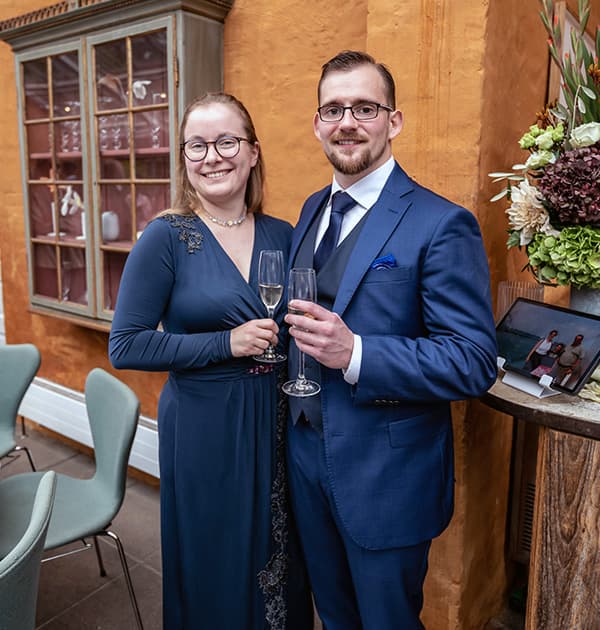 Andrea Plovgaard Frederiksen and her partner dressed formally and smiling while holding champagne glasses at a celebration.
