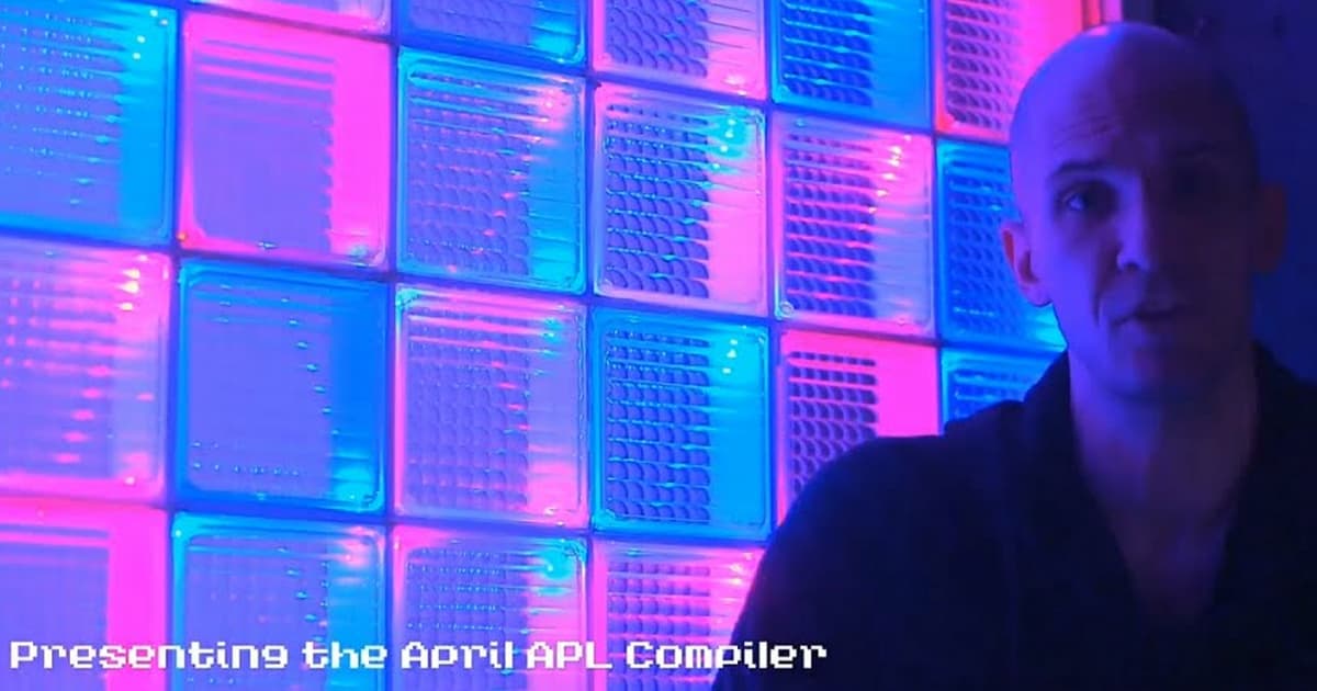 Andrew Sengul presenting April: An APL Compiling to Common Lisp at APL Seeds ’22, standing beside an illuminated glass-block wall glowing in blue and pink tones.