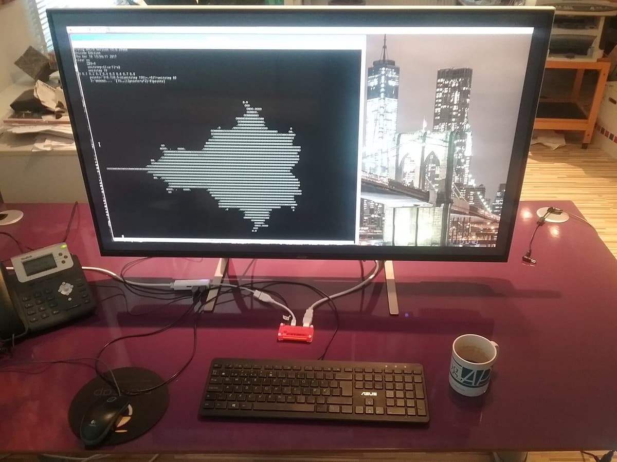 Desktop setup with a large monitor displaying a text-mode Mandelbrot in Dyalog APL; keyboard, mouse and cables on a purple desk, with a small red single-board computer and an APL mug.