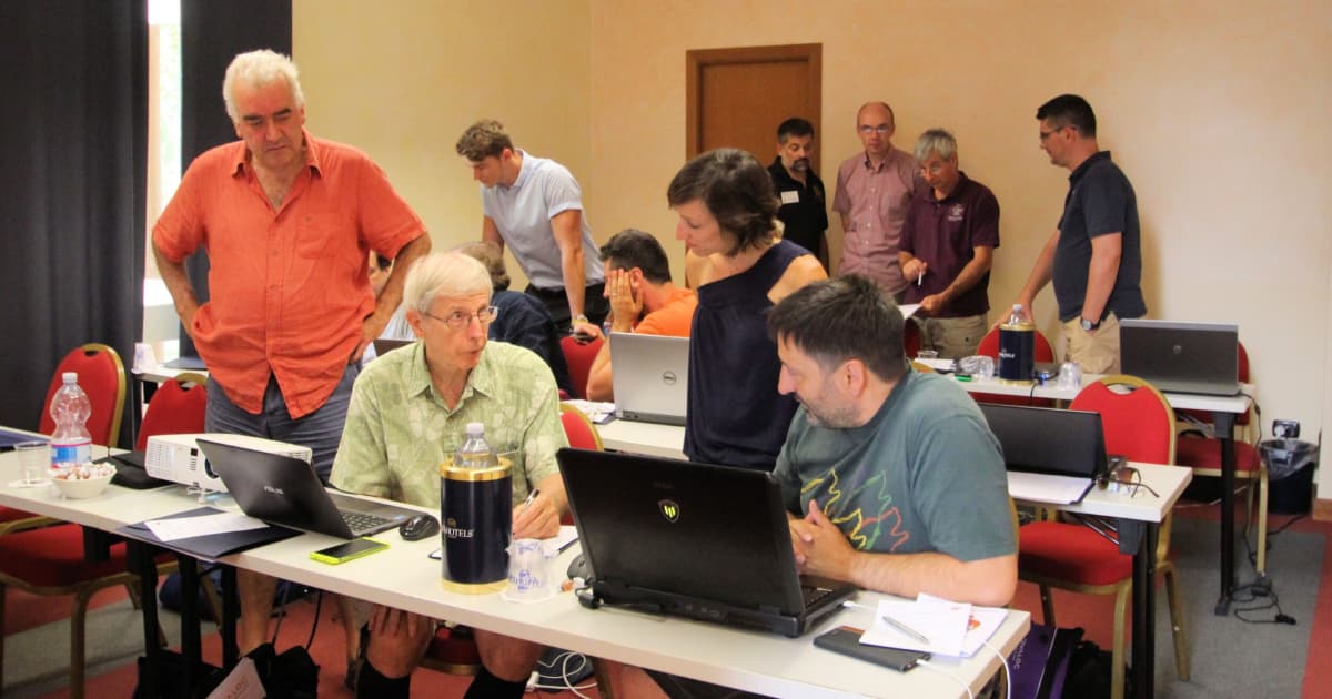 Workshop classroom with attendees collaborating at tables, discussing code and working on laptops during a Dyalog MiServer web-app session.