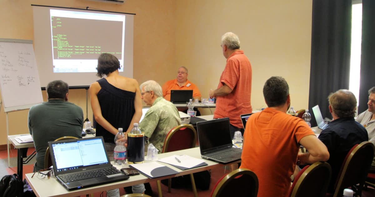 Workshop room with attendees seated at tables watching a projected code demo, presenter at the front, laptops and a flipchart visible.