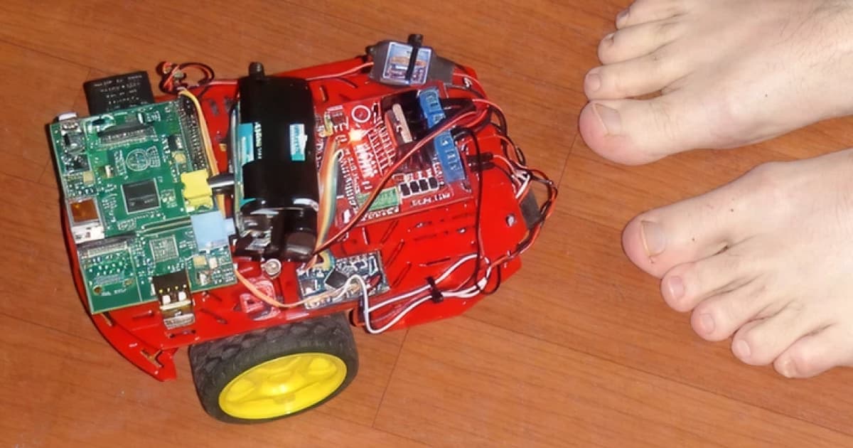 Top-down view of a small red C3Pi Raspberry Pi robot with exposed electronics, beside a pair of bare feet on a wooden floor