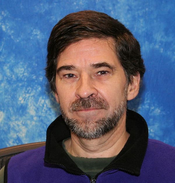 Dan Baronet seated in front of a mottled blue backdrop, wearing a fleece jacket and looking into the camera with a calm expression.