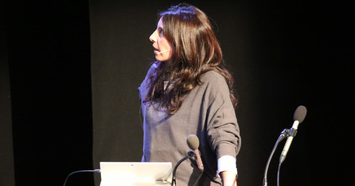 Arianna Francia from SimCorp Italiana presenting on stage at the Dyalog ’18 user meeting in Belfast, Northern Ireland. She is wearing a grey top with rolled-up sleeves and a headset microphone, standing beside a lectern with a laptop and microphones, looking towards the projection screen.