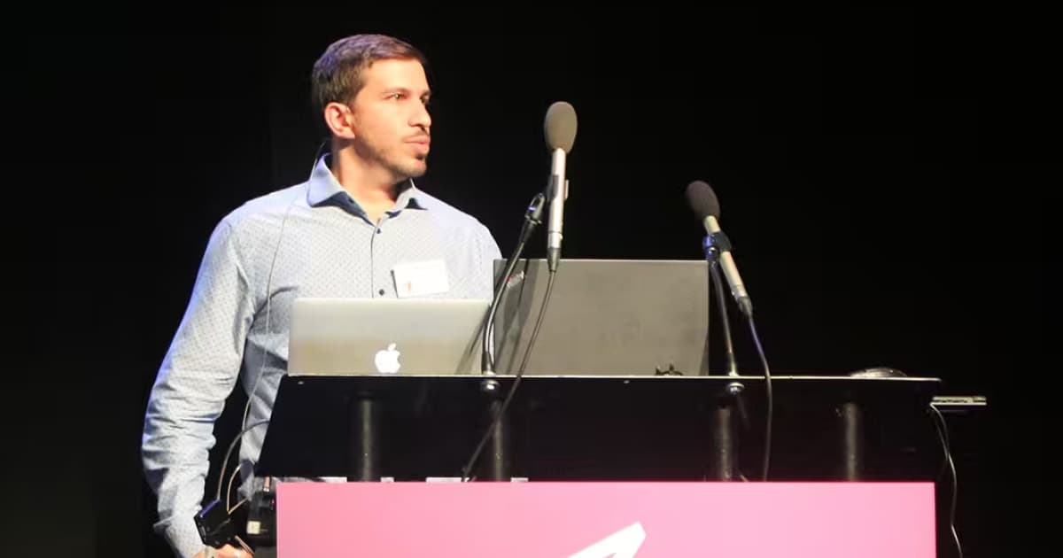 Gilgamesh Athoraya presenting on stage at the Dyalog ’18 user meeting in Belfast, Northern Ireland. He is wearing a light patterned shirt and speaking into a headset microphone, standing behind a lectern with two laptops and several microphones.