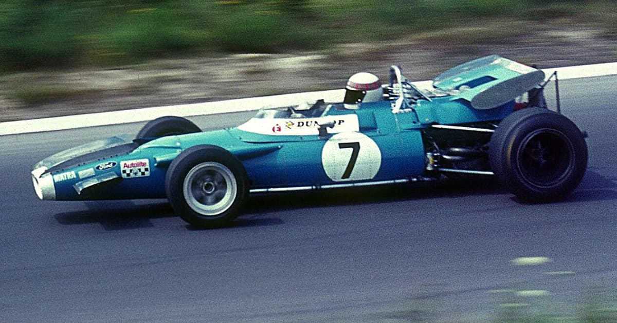A vintage Formula One race car in teal with the number 7 on its side, photographed in motion on a racetrack. The driver is wearing a red and white helmet, and sponsor logos including Dunlop, Ford, and Autolite are visible on the car’s bodywork.