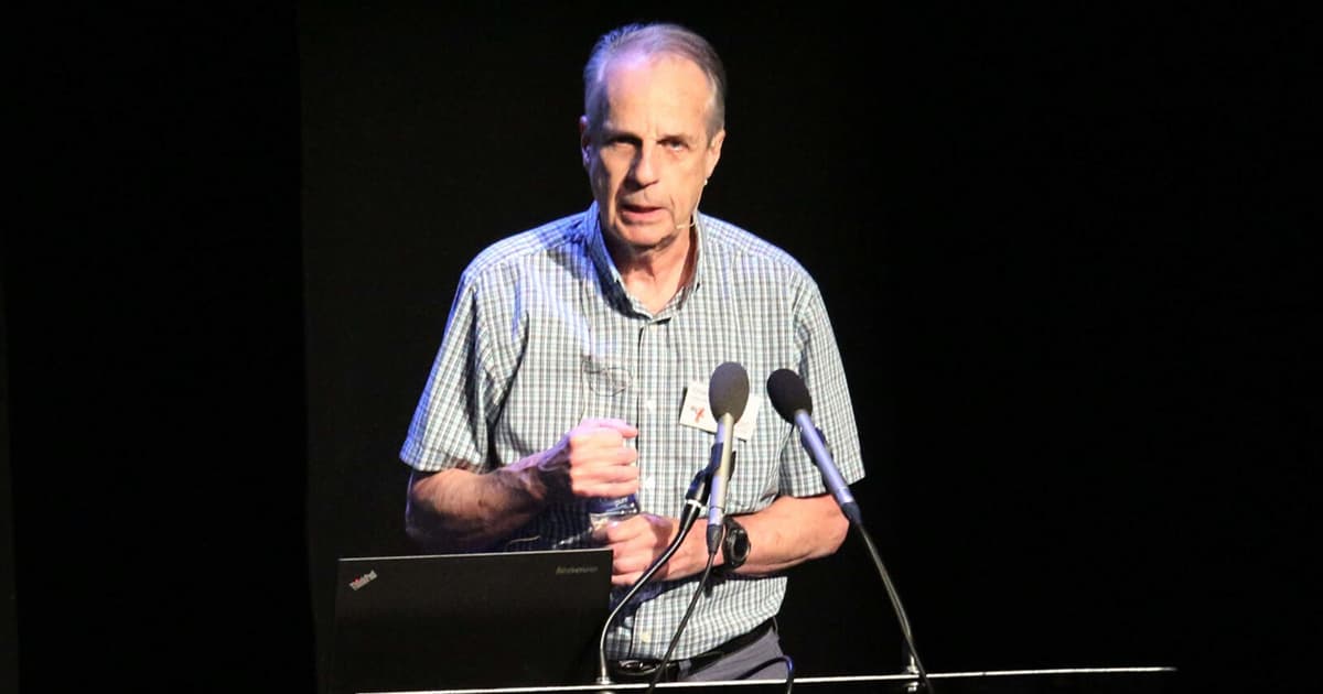Charles Brenner presenting on stage at the Dyalog ’18 user meeting in Belfast, Northern Ireland. He is wearing a green and white checked short-sleeve shirt with a name badge and headset microphone, standing behind a lectern with a laptop and two microphones, holding a water bottle.