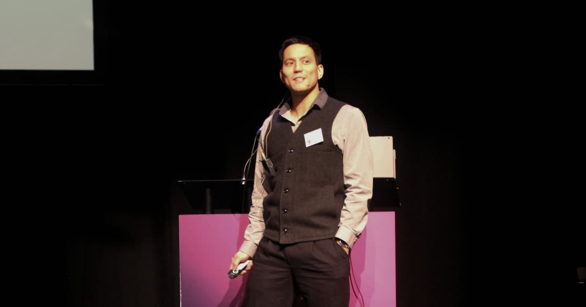 Aaron Hsu speaking at the Dyalog ’18 user meeting in Belfast, Northern Ireland. He is standing on stage wearing a dark waistcoat over a light shirt, holding a presentation remote, with a microphone headset.