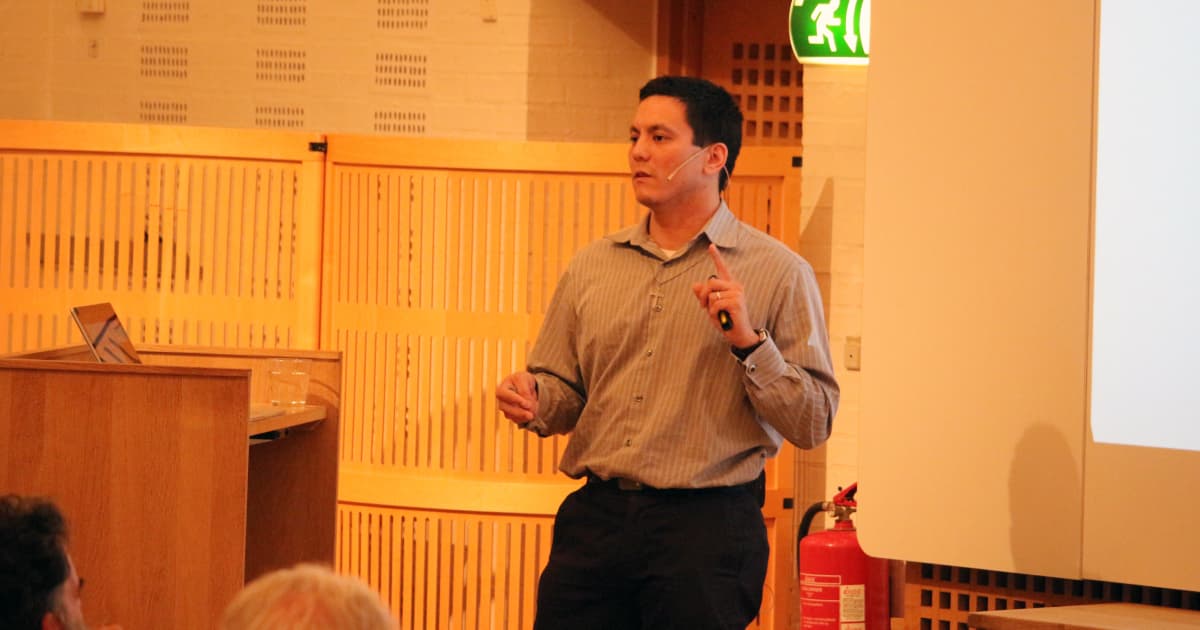 Aaron Hsu presenting at Dyalog ’19 in Elsinore, standing beside the lectern and gesturing with a presentation remote, wearing a grey shirt and headset microphone.
