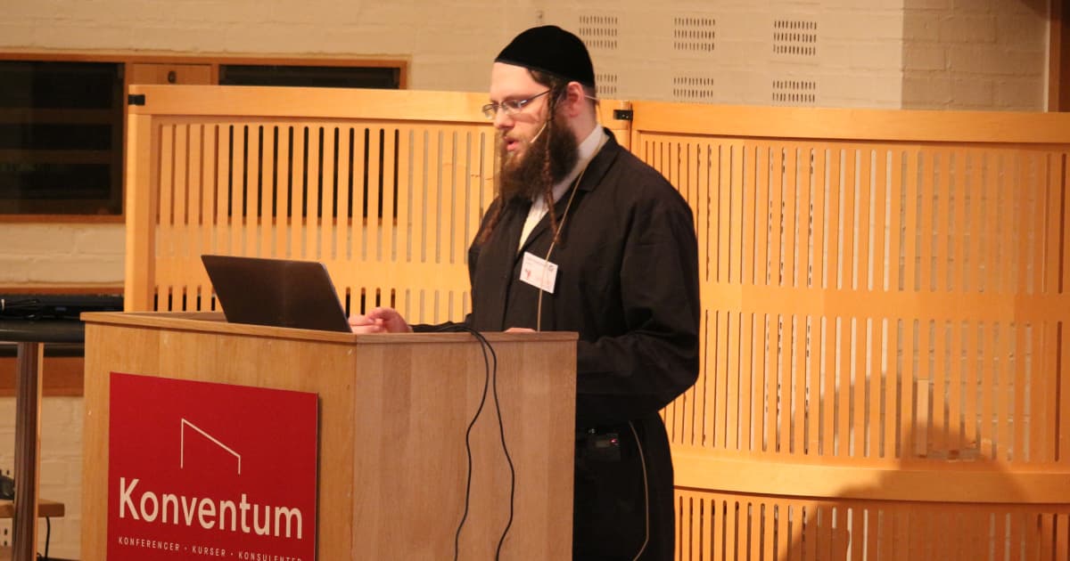 Adám Brudzewsky presenting at Dyalog ’19 in Elsinore. He stands at a wooden podium with a laptop, speaking into a headset microphone. The podium has a red sign reading “Konventum.