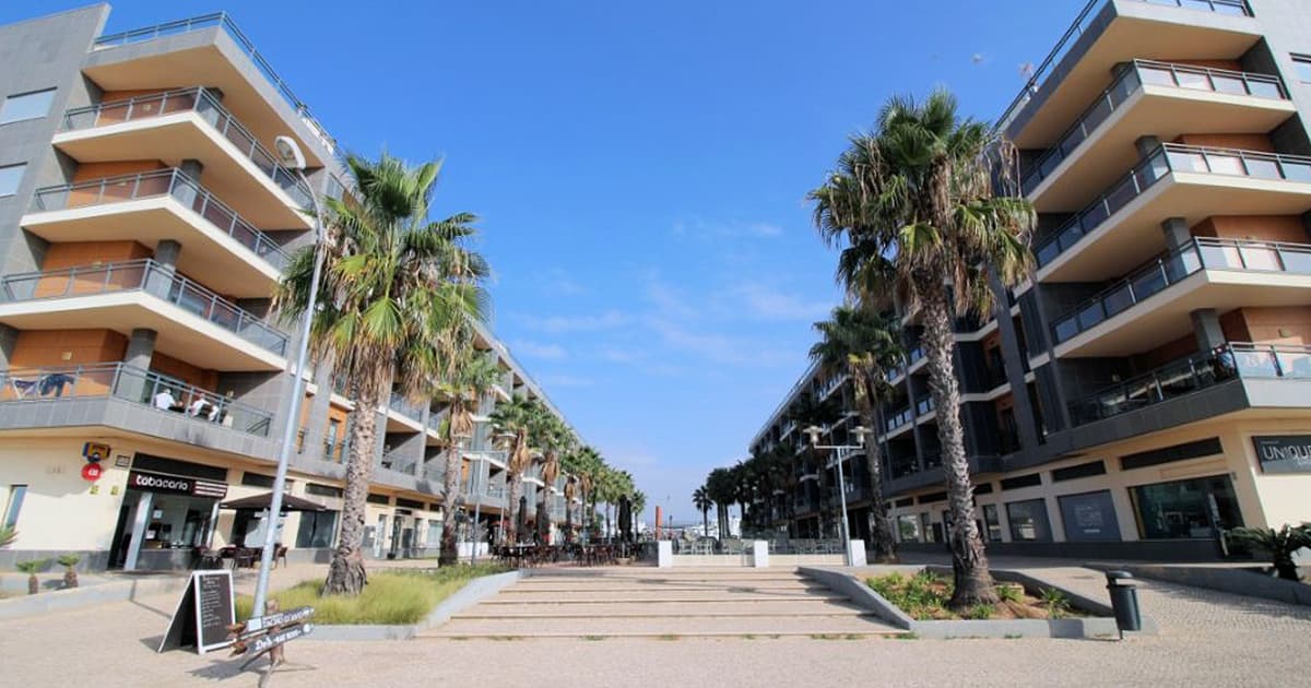 View of the central promenade at Marina Village in Olhão, Portugal, flanked by modern apartment buildings with palm trees lining the walkway under a clear blue sky.