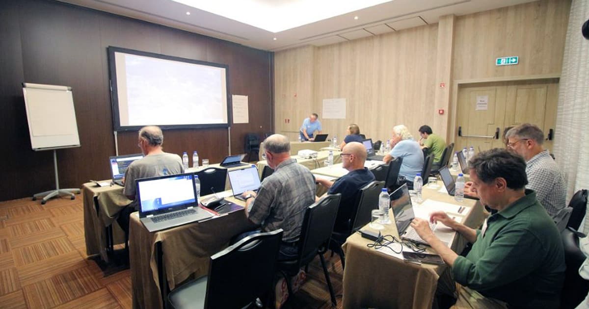Attendees working on laptops during a workshop at Dyalog '22, seated at tables with water bottles and notebooks in a conference room with a large screen at the front.