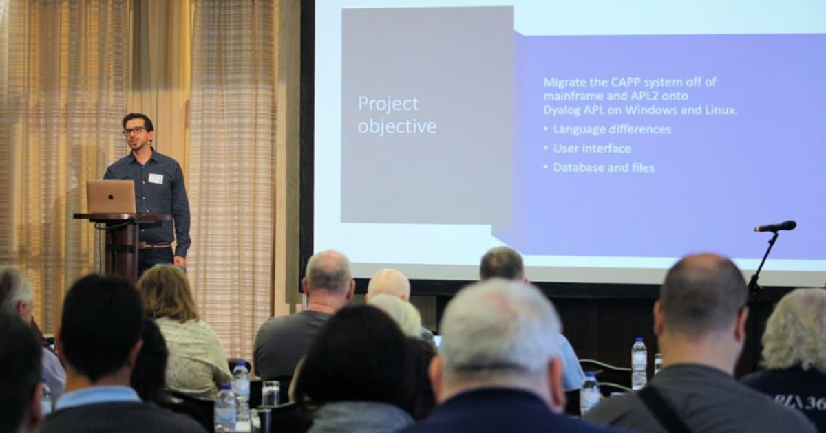 Gilgamesh Athoraya presenting at Dyalog '22 in Olhão, Portugal, discussing the migration of an APL2 mainframe system to Dyalog APL in the cloud, with a slide showing project objectives projected behind him.