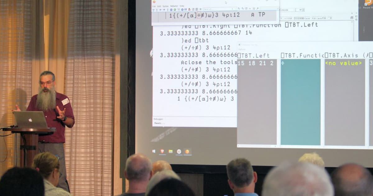 John Daintree presenting at Dyalog '22 in Olhão, Portugal, demonstrating inline “token by token” debugging in APL with a live session projected on screen behind him.