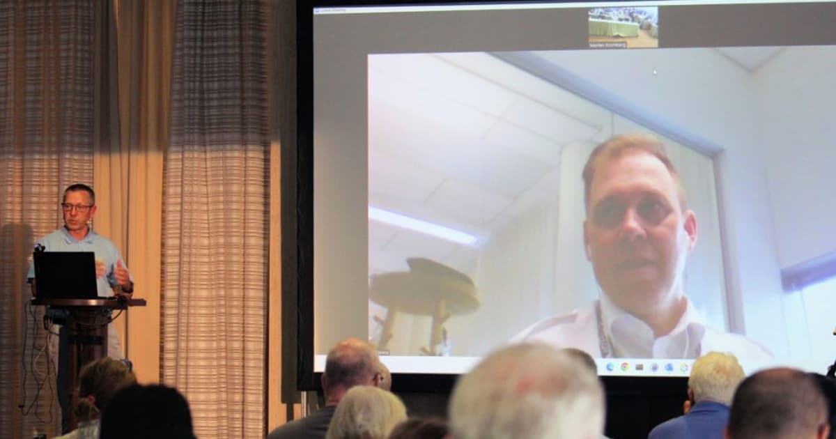 Morten Kromberg standing at a podium during Dyalog '22 in Olhão, Portugal, while Kimmo Linna appears on a large screen via video call, presenting his workflow integrating Dyalog, DuckDB, Vega-lite, and Jupyter.