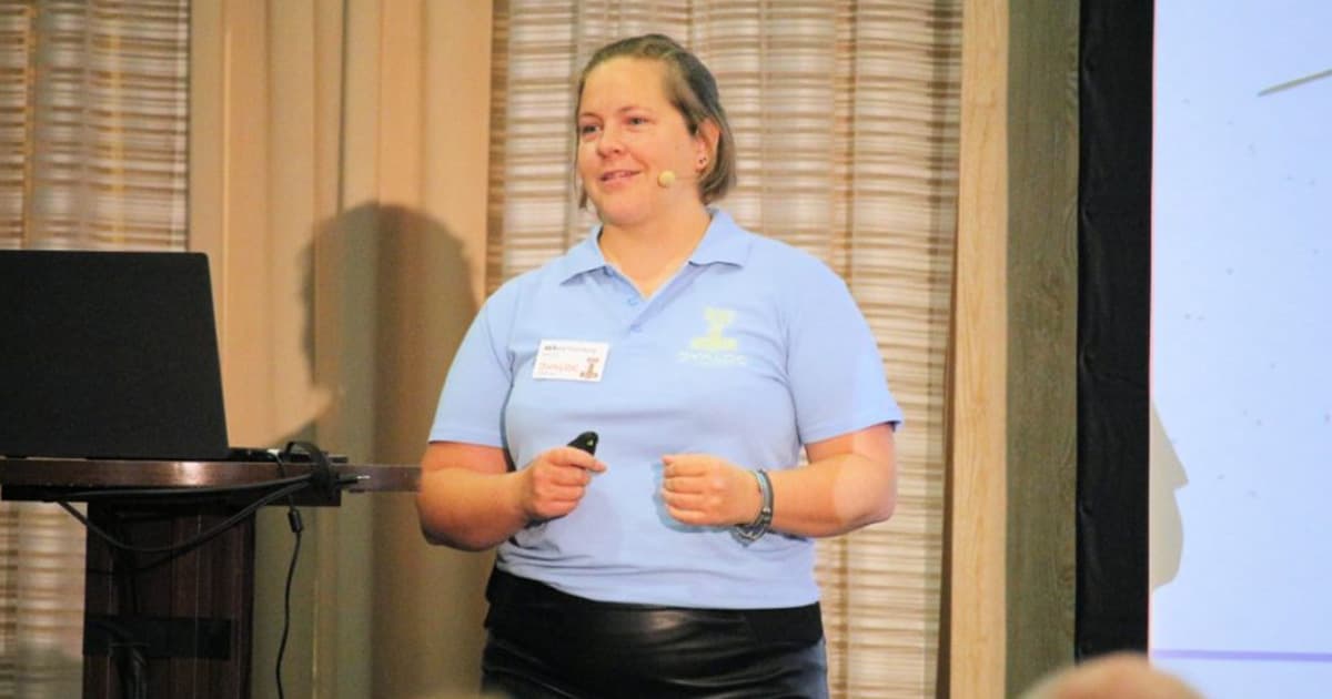 Stine Kromberg speaking at Dyalog '22 in Olhão, Portugal, smiling as she presents while standing beside a podium with a laptop, wearing a light blue Dyalog shirt and a headset microphone.