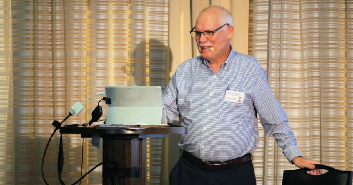 Stephen Mansour presenting at Dyalog '22 in Olhão, Portugal, explaining the TAMPA mathematical programming framework while standing at a podium with a headset microphone and a tablet.