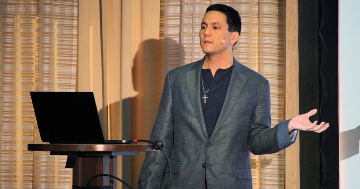 Aaron Hsu standing on stage at Dyalog '22, wearing a grey blazer and dark shirt with a cross necklace, speaking into a headset microphone. He gestures with one hand while holding a clicker in the other. A laptop on a podium and a large projection screen are beside him, with curtains in the background.