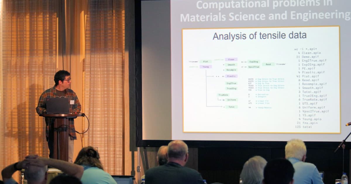 Jesús Galán López stands at a podium with a laptop, giving a presentation at Dyalog ’22. The slide on the large screen is titled “Computational problems in Materials Science and Engineering” and shows an “Analysis of tensile data” flow diagram with code files and functions related to data processing. The audience is seated at tables, watching the talk.