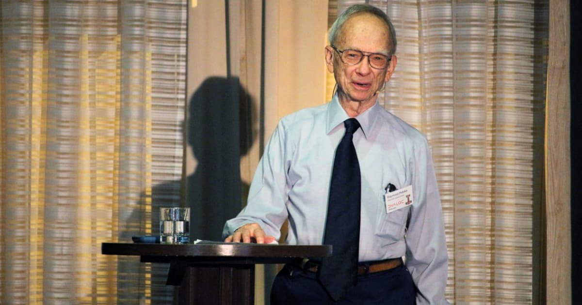Ray Polivka stands at a podium during Dyalog ’22, wearing a light blue shirt and dark tie. He is speaking to the audience while introducing his new book for high school students. A glass of water sits on the podium, and patterned curtains form the backdrop.