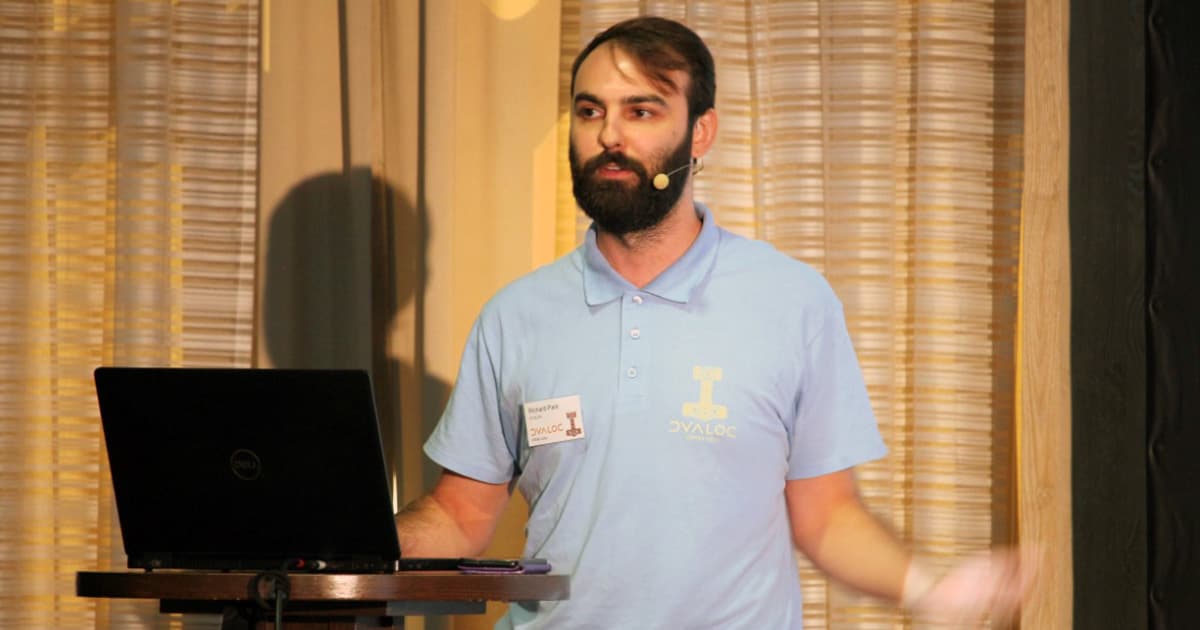 Rich Park speaks at Dyalog ’22, standing beside a laptop on a podium. He wears a light blue Dyalog polo shirt and headset microphone. Rich gestures with his right hand while addressing the audience about media, training, and outreach.