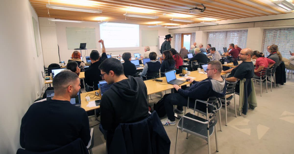 Participants seated with laptops during a workshop at Dyalog ’23 in Denmark, with one attendee raising a hand and Adám Brudzewsky standing near the centre of the room.