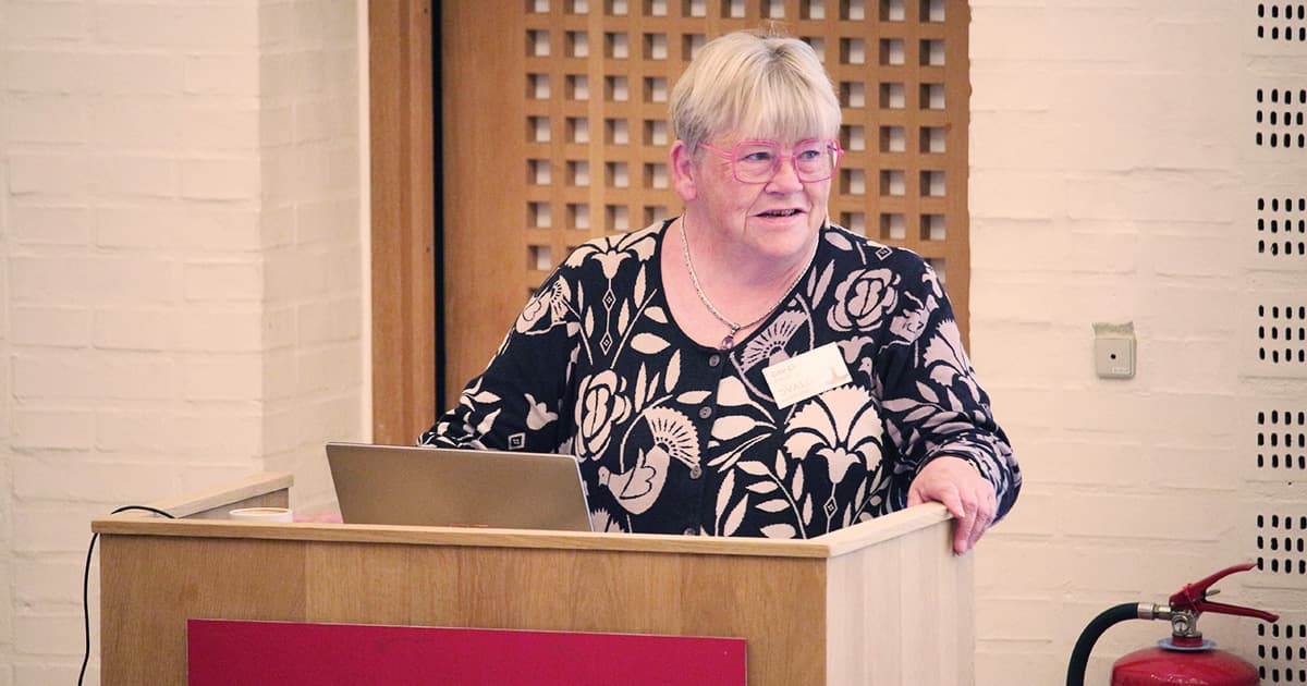 Gitte Christensen speaking from a podium with a laptop during Dyalog ’23, wearing a patterned black and white top and pink glasses.