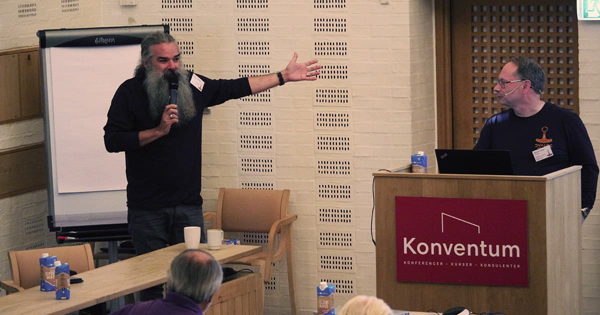 John Daintree gestures while speaking into a microphone beside a flipchart, as Richard Smith stands at the podium with a laptop during their joint presentation at Dyalog ’23 in Konventum.