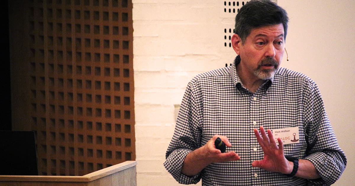 Mark Wolfson speaking at Dyalog ’23, standing at a podium with a microphone headset, gesturing with one hand while holding a remote in the other.