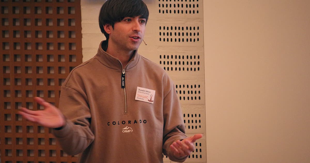 Brandon Wilson speaking at Dyalog ’23, gesturing with both hands while presenting in front of a light-coloured wall.