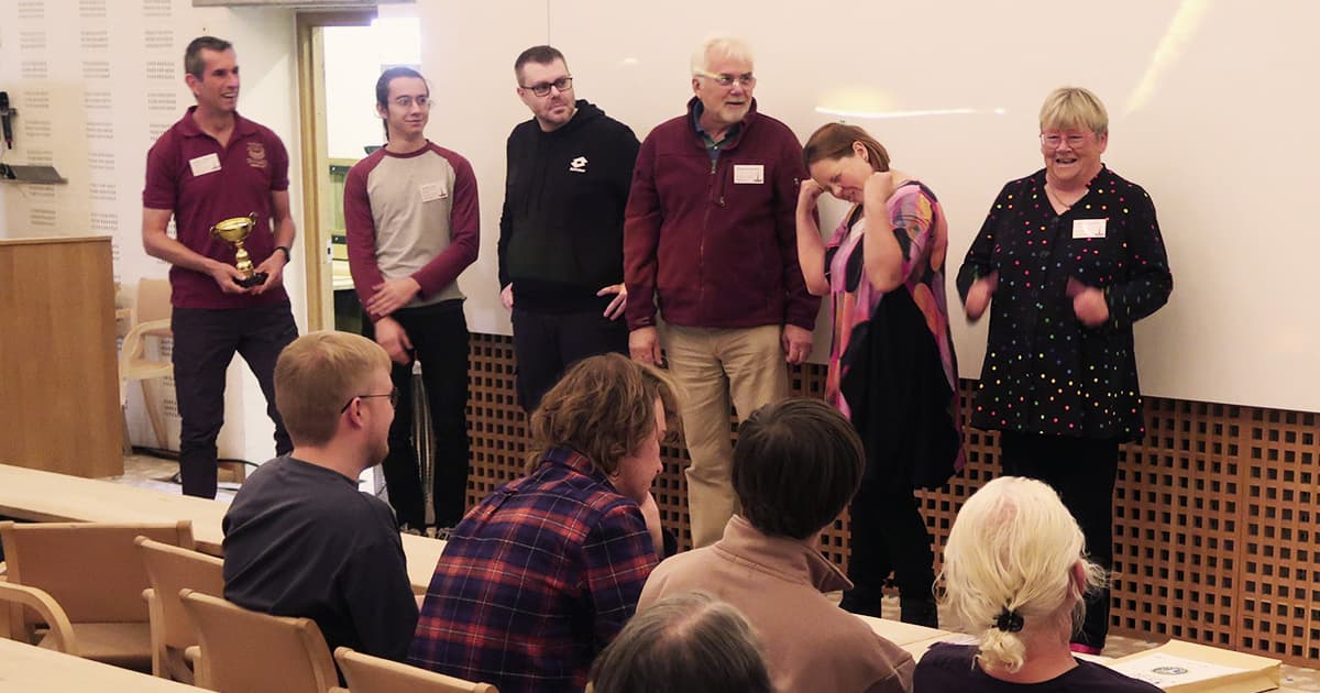 The winning team of the Viking Challenge at Dyalog ’23 stands at the front of the room with a trophy, smiling and celebrating, while the audience applauds.