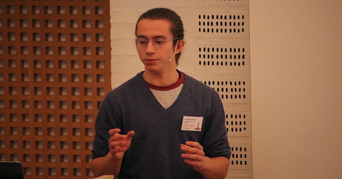 Andrea Piseri presenting at Dyalog ’23, wearing glasses and a dark jumper, speaking with both hands raised slightly while addressing the audience.