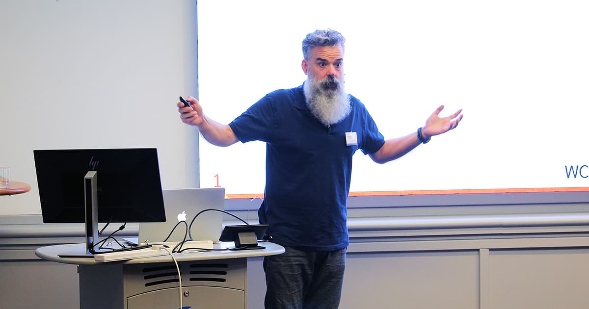 John Daintree stands at the front of a lecture room at Dyalog ’24, gesturing with both arms while presenting slides on new functionality. A computer and projector screen are set up beside him.