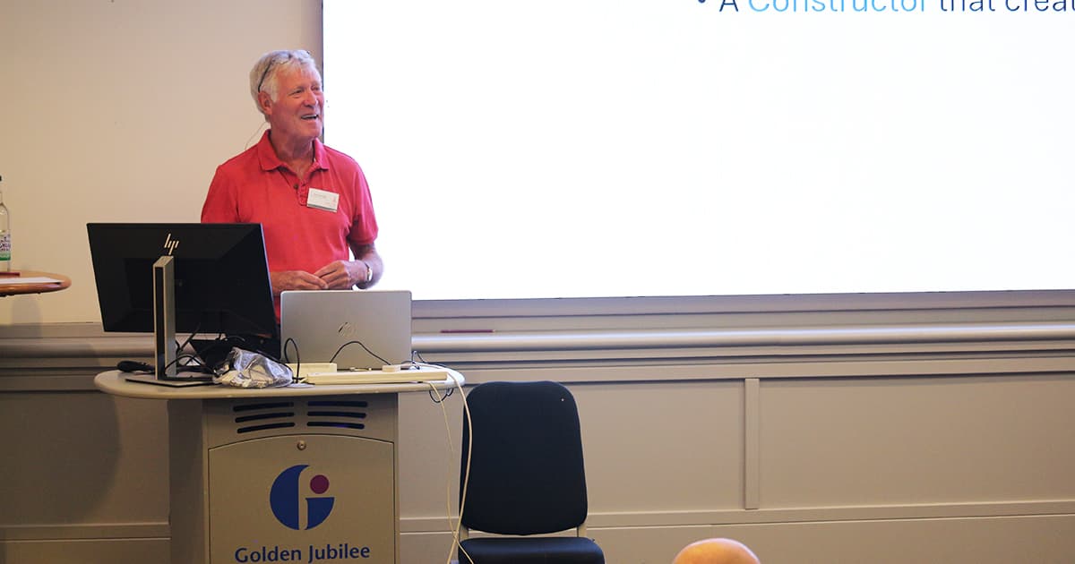 Pete Donnelly presenting at Dyalog ’24 in Glasgow, standing at the lectern with a laptop in front of him and speaking in front of a projected slide. He is wearing a red shirt.