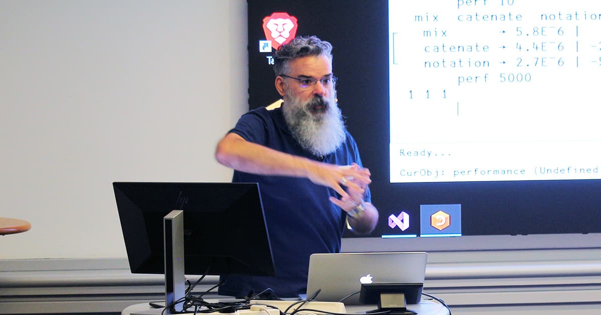 John Daintree presenting at Dyalog ’24 in Glasgow. He is standing beside a lectern with a computer, gesturing with both hands, while slides showing APL code and performance results are projected behind him.