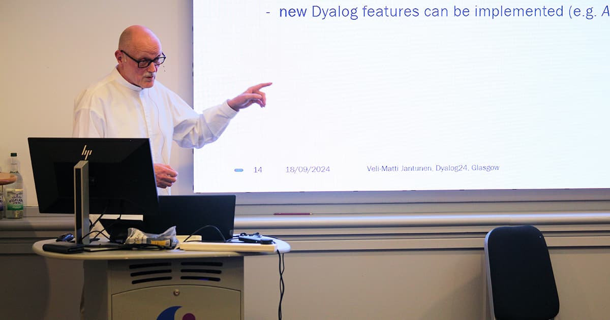 Veli-Matti Jantunen presenting at Dyalog ’24 in Glasgow. He is standing at a lectern, pointing towards a projected slide that discusses implementing new Dyalog features, with his name and the event details visible on the slide.