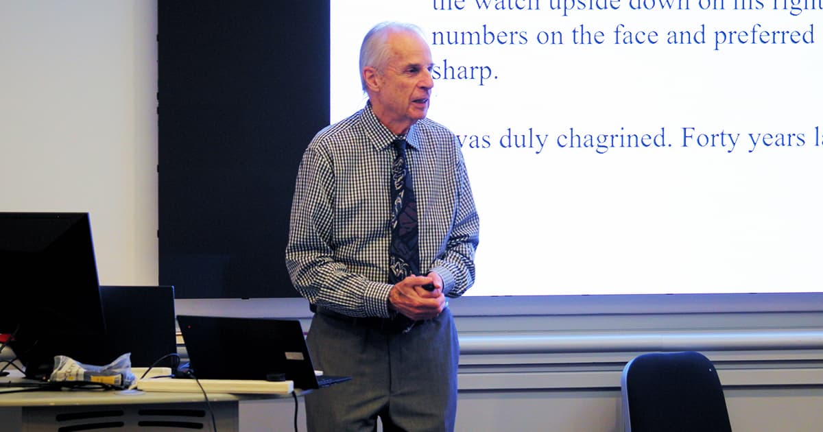 Charles Brenner speaking at Dyalog ’24 in Glasgow. He is standing at the front of the room in a checked shirt and tie, delivering his presentation “Some APL Pioneers I Knew” with projected text visible behind him.