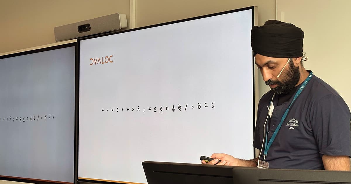 Karta Kooner presenting at PLSS 2025 in Bergen, Norway. He stands beside two large screens displaying APL glyphs, with the Dyalog logo shown in the top-left corner.