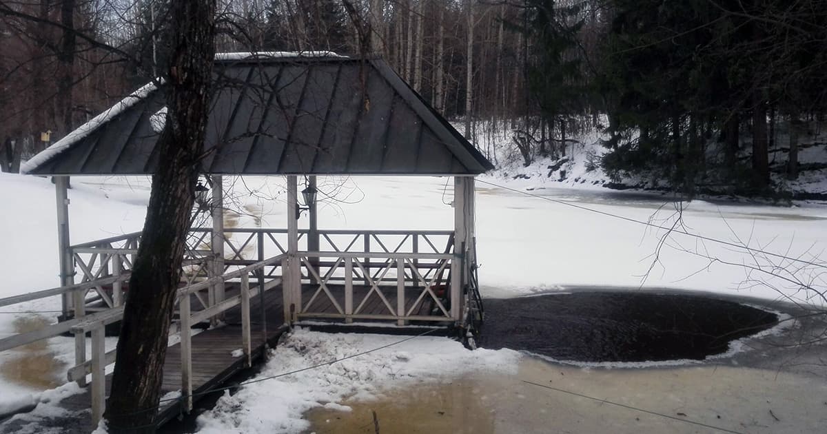 A snow-covered wooden gazebo with a ramp extending to the edge of a partially frozen pond, surrounded by a winter forest. An ice hole near the gazebo suggests it was used for cold-water immersion.