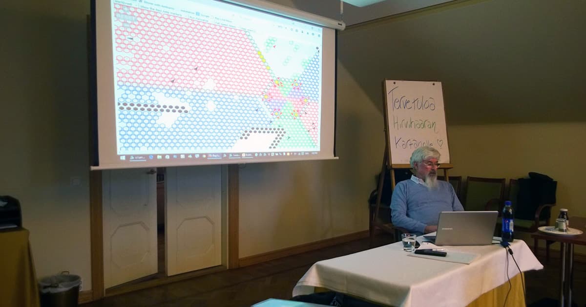 Ray Cannon sits beside a projection screen displaying a colourful hexagonal grid, likely part of a simulation involving ants. A whiteboard in the background welcomes participants in Finnish to the FinnAPL Forest Seminar.
