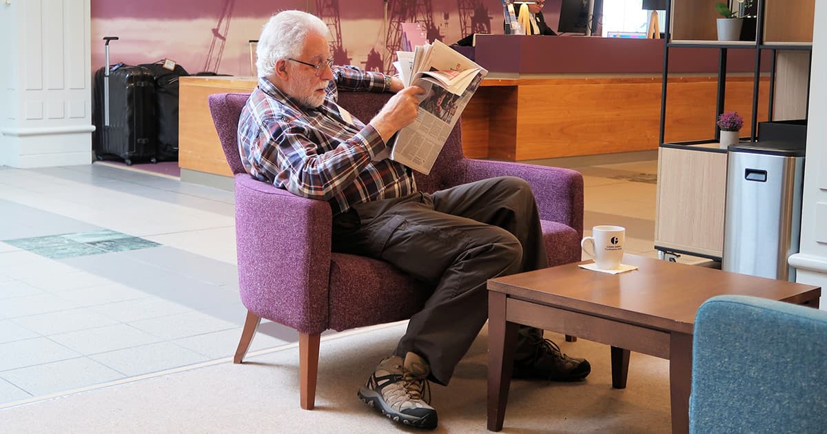 Geoff Streeter seated in the reception area of the Golden Jubilee Hotel in Glasgow, Scotland, during the Dyalog '24 user meeting, reading a newspaper with a coffee cup on the table beside him.