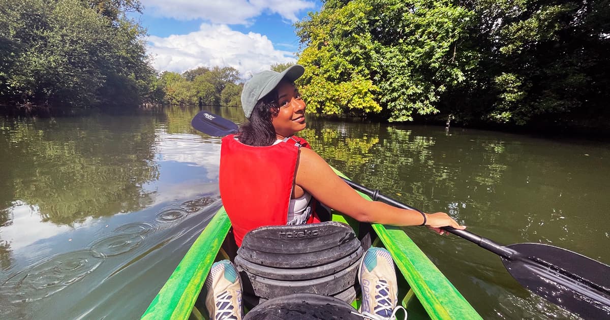 Jada Andrade kayaking on a calm river, smiling back at the camera while wearing a red life jacket and green cap.